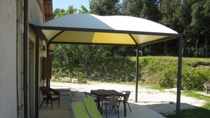 Ein überdachter Außenbereich mit Tischen und Stühlen, eine Pergola spendet Schatten, und Blick auf eine natürliche Landschaft.