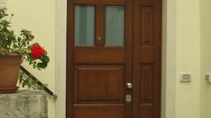 Una puerta de madera con un ventanal, una maceta con flores rojas y un muro de piedra.