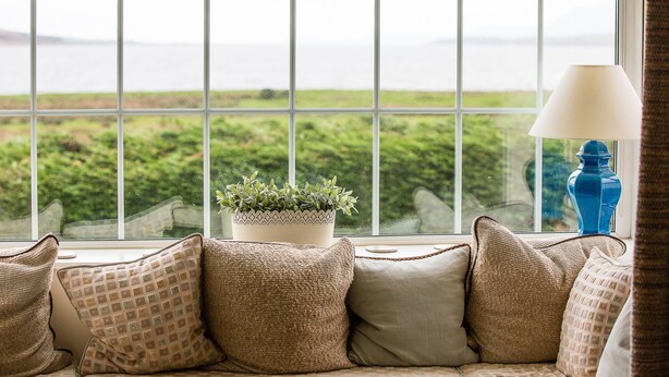Una ventana con vistas a zonas verdes y al agua, un sofá con cojines estampados, y un florero azul en el alféizar.