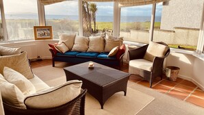 Una habitación iluminada por el sol con sofá, sillas y una mesa de centro. Amplias ventanas ofrecen vista al mar y al verdor.