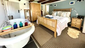 A bedroom with a wooden bed, a nightstand, a lamp, a bedside table, a large window, and a bathroom with a bathtub and toiletries.