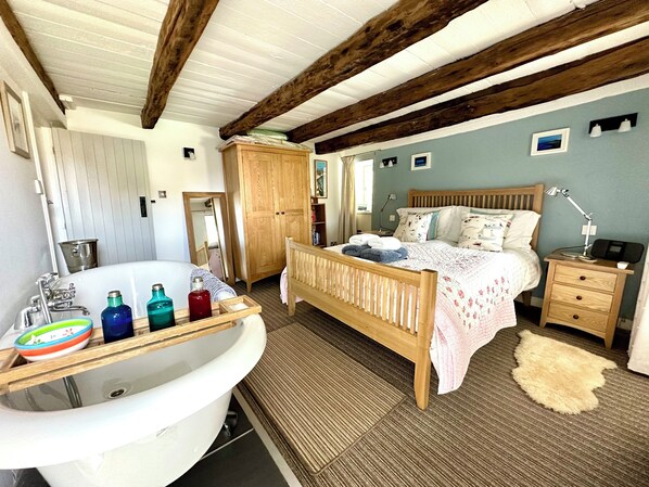 Une chambre avec un lit en bois, une table de chevet, une lampe, une commode, une grande fenêtre et une salle de bain avec une baignoire et des articles de toilette.