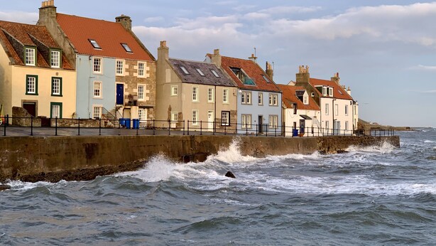 A coastal town with buildings along the waterfront, a stone sea wall, and waves crashing against it.
