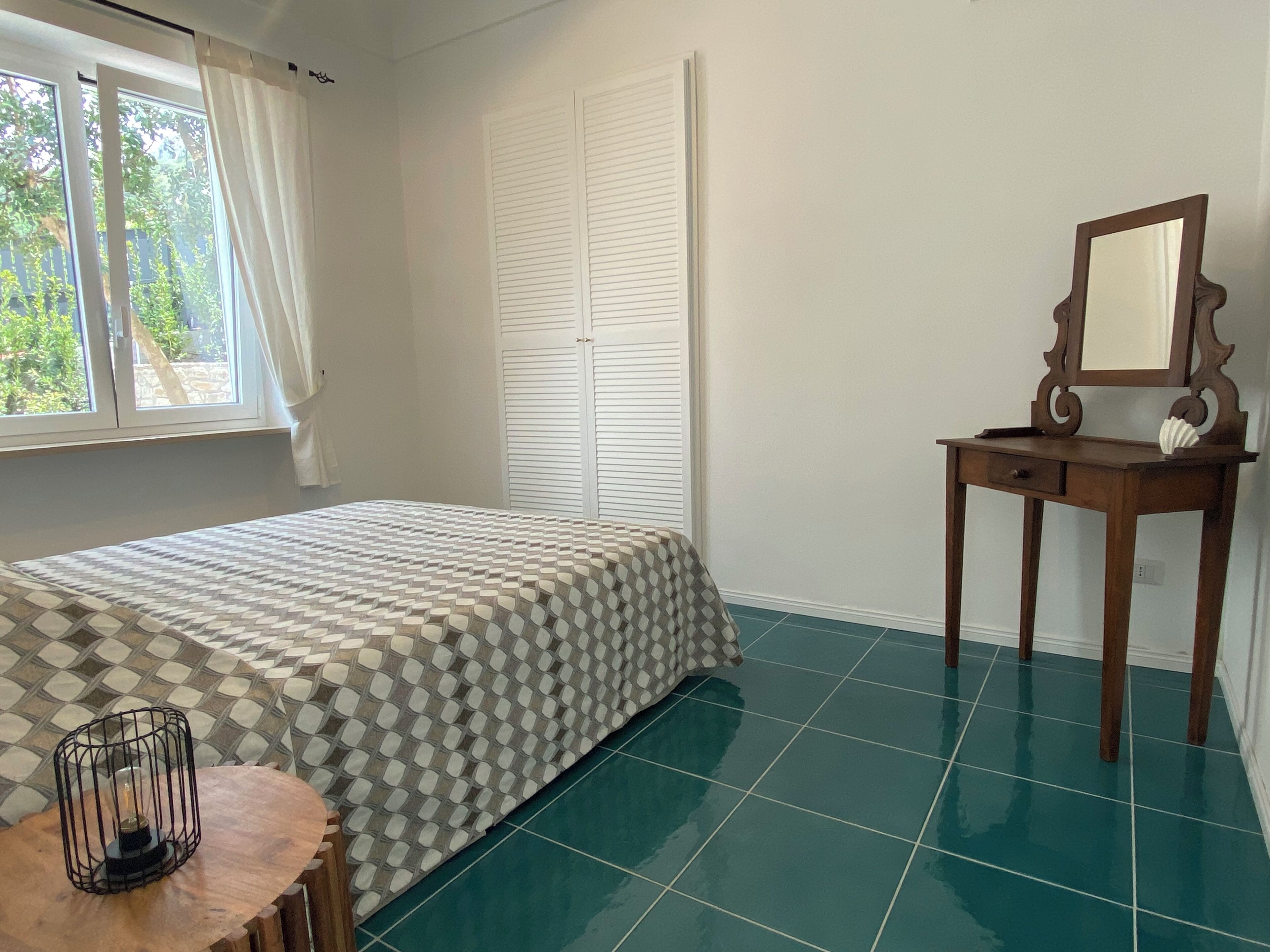 A bedroom with a bed, a bedside table with a mirror, a small table with a candle, and a window with curtains.