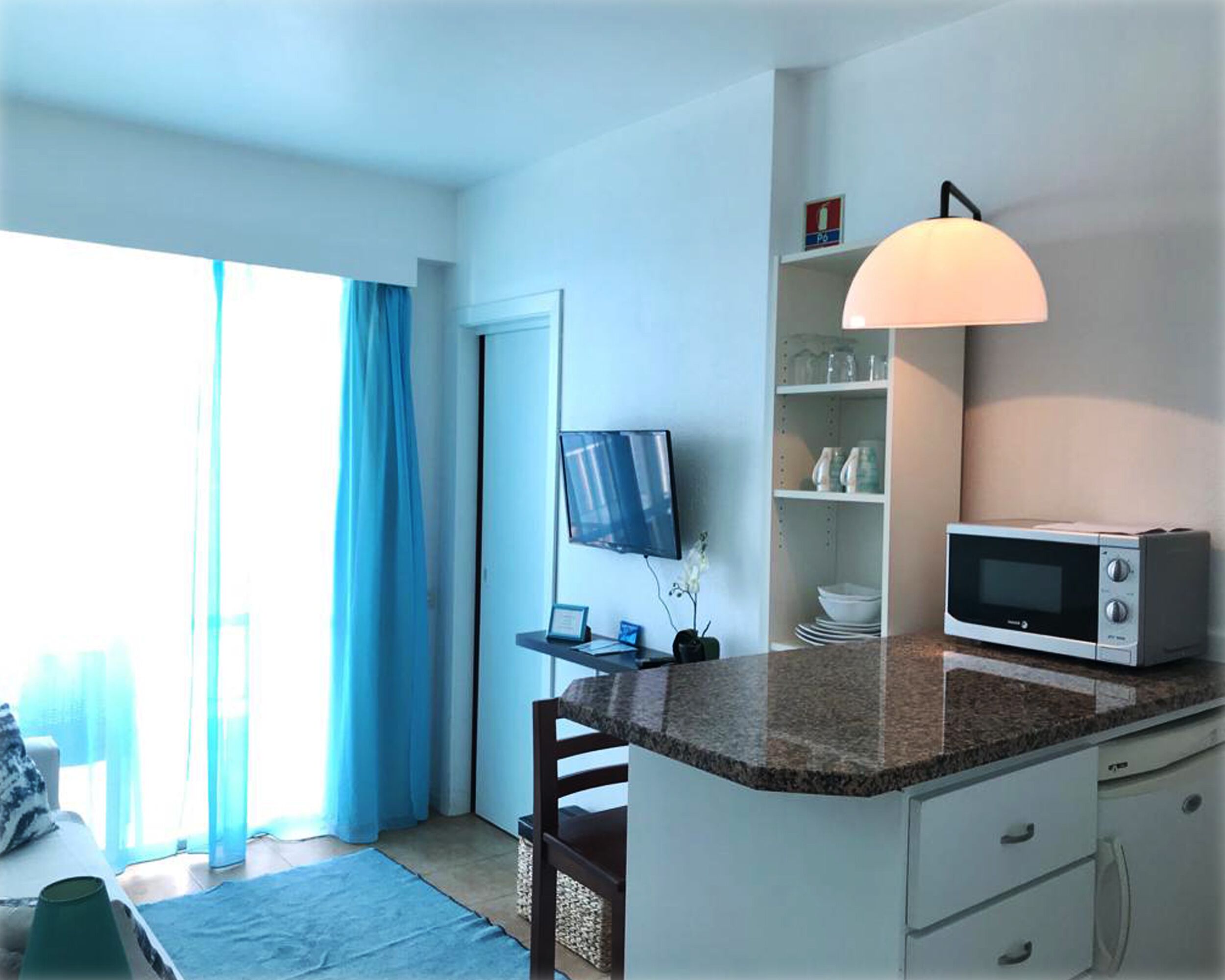 A compact kitchen with a microwave, a small shelf with dishes, and a TV mounted on the wall.