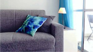 A grey sofa with a blue and green patterned pillow, a floor lamp, and a view of the outdoors through a window with blue curtains. - Bright Seafront Apartment In Funchal with Sea View from the Balcony (Funchal)