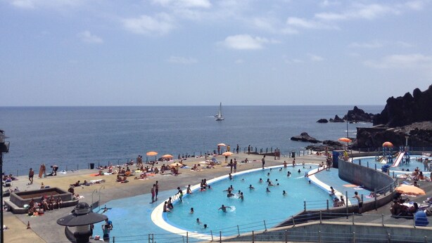 A beachside swimming pool with people sunbathing and swimming, a sailboat in the distance, and rocky cliffs. - Bright Seafront Apartment In Funchal with Sea View from the Balcony (Funchal)