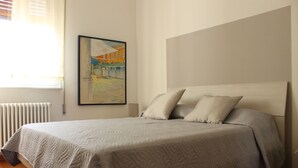 A neatly made bed with a quilted cover, a framed painting on the wall, and a radiator beside the window.