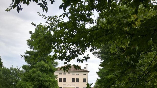 A serene pond with a reflection of a white building with multiple windows and a symmetrical facade, surrounded by lush greenery and trees.