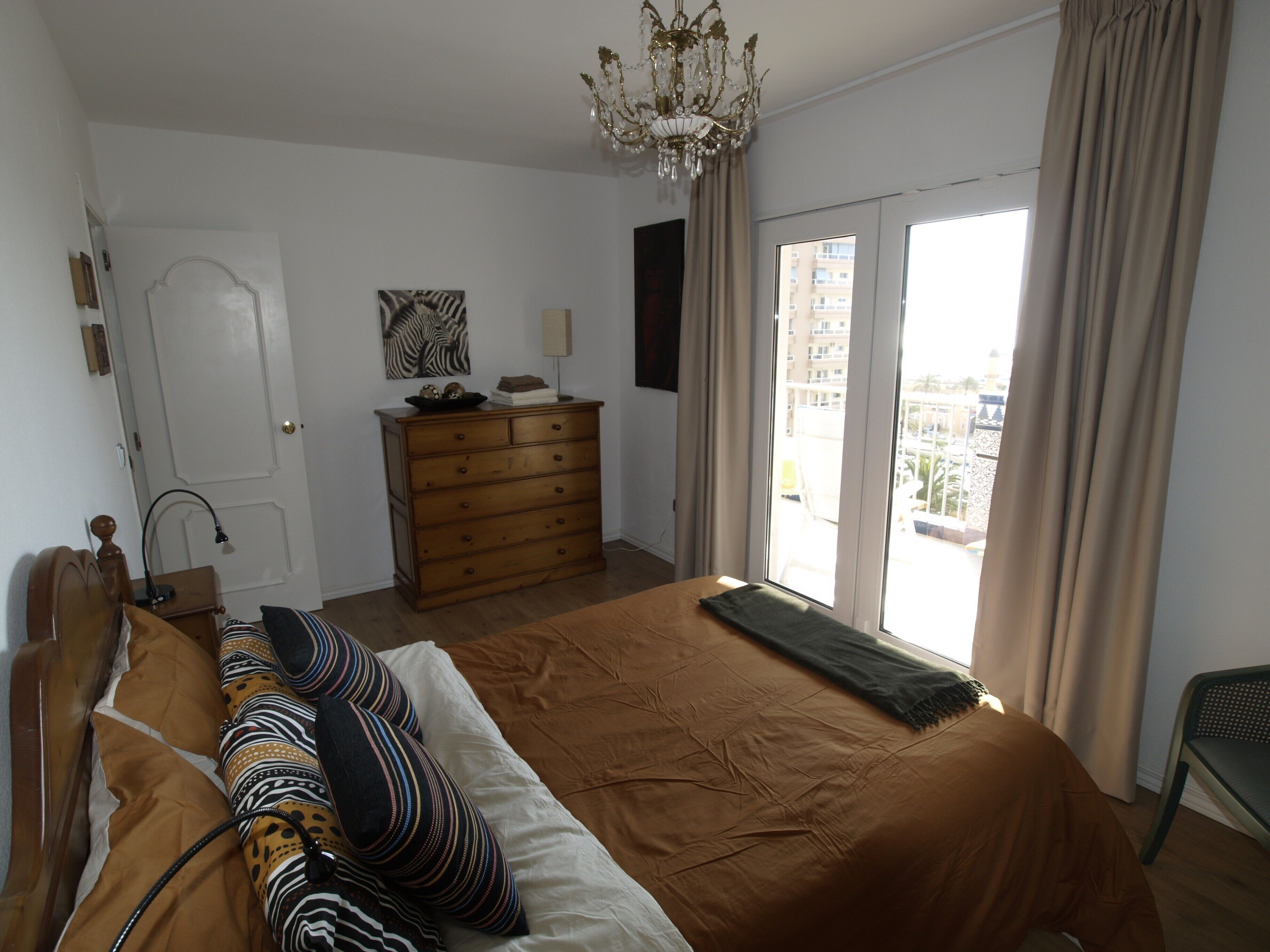 A bedroom with a bed, a dresser, a chandelier, a door, and a window with curtains.