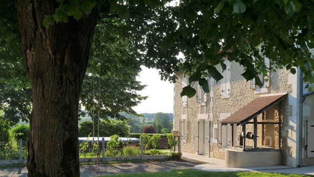Un edificio con facciata in pietra, un grande albero in primo piano e una zona verde con altalene.
