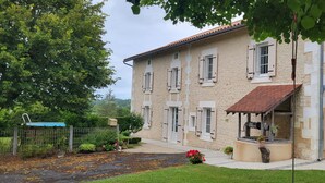 Un edificio a due piani con facciata in pietra e tetto in tegole rosse. È presente un portico coperto con una panca in legno e piante in vaso.
