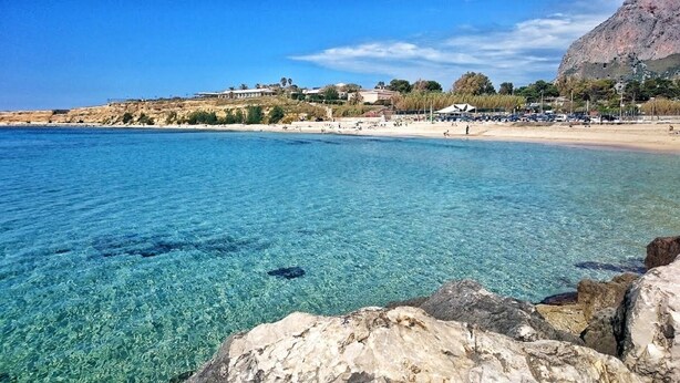 A coastal beach with clear blue water, rocky shoreline, and buildings in the background.