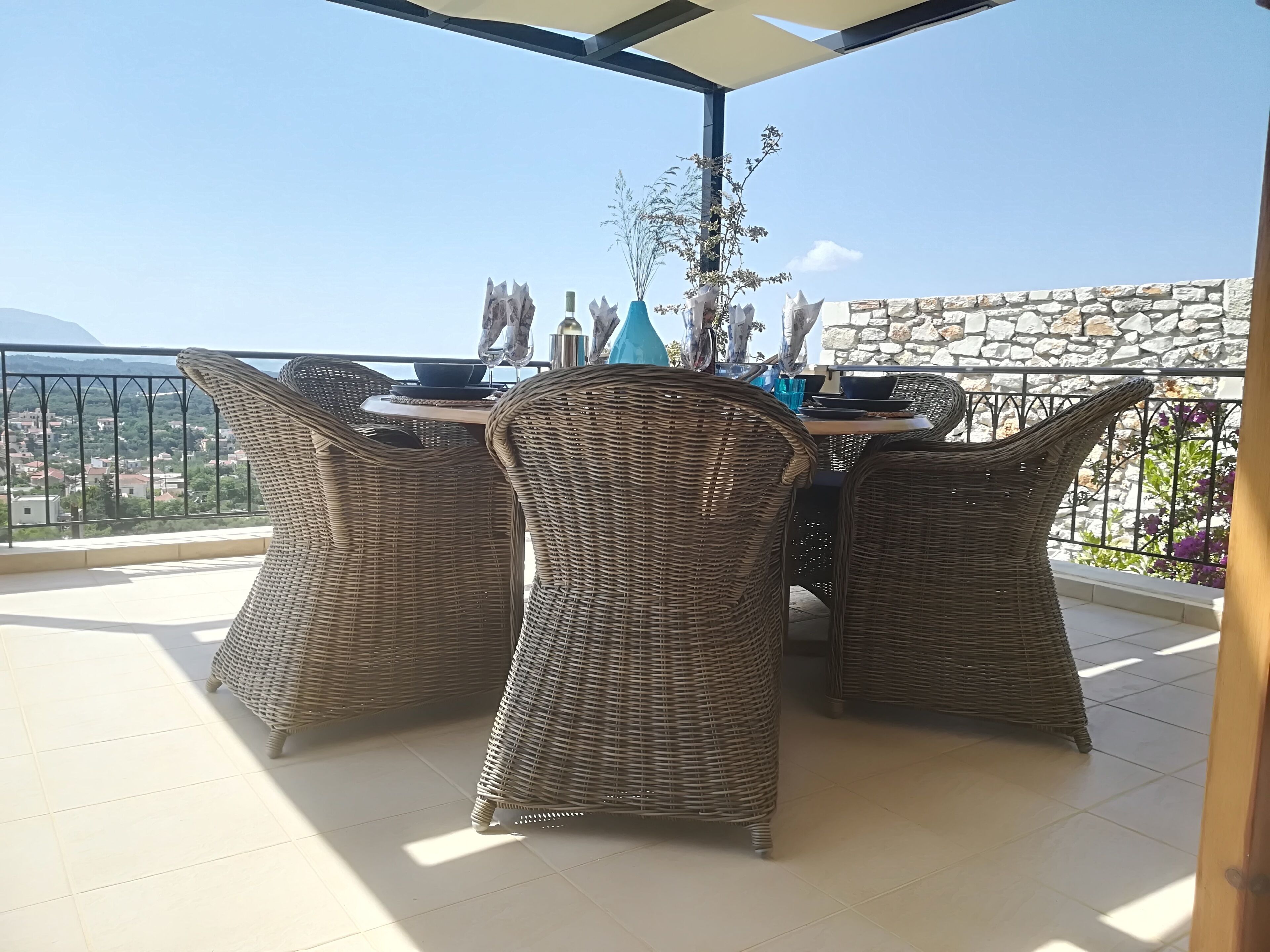 A covered outdoor dining area with wicker chairs and a table, overlooking a scenic view of houses and mountains.