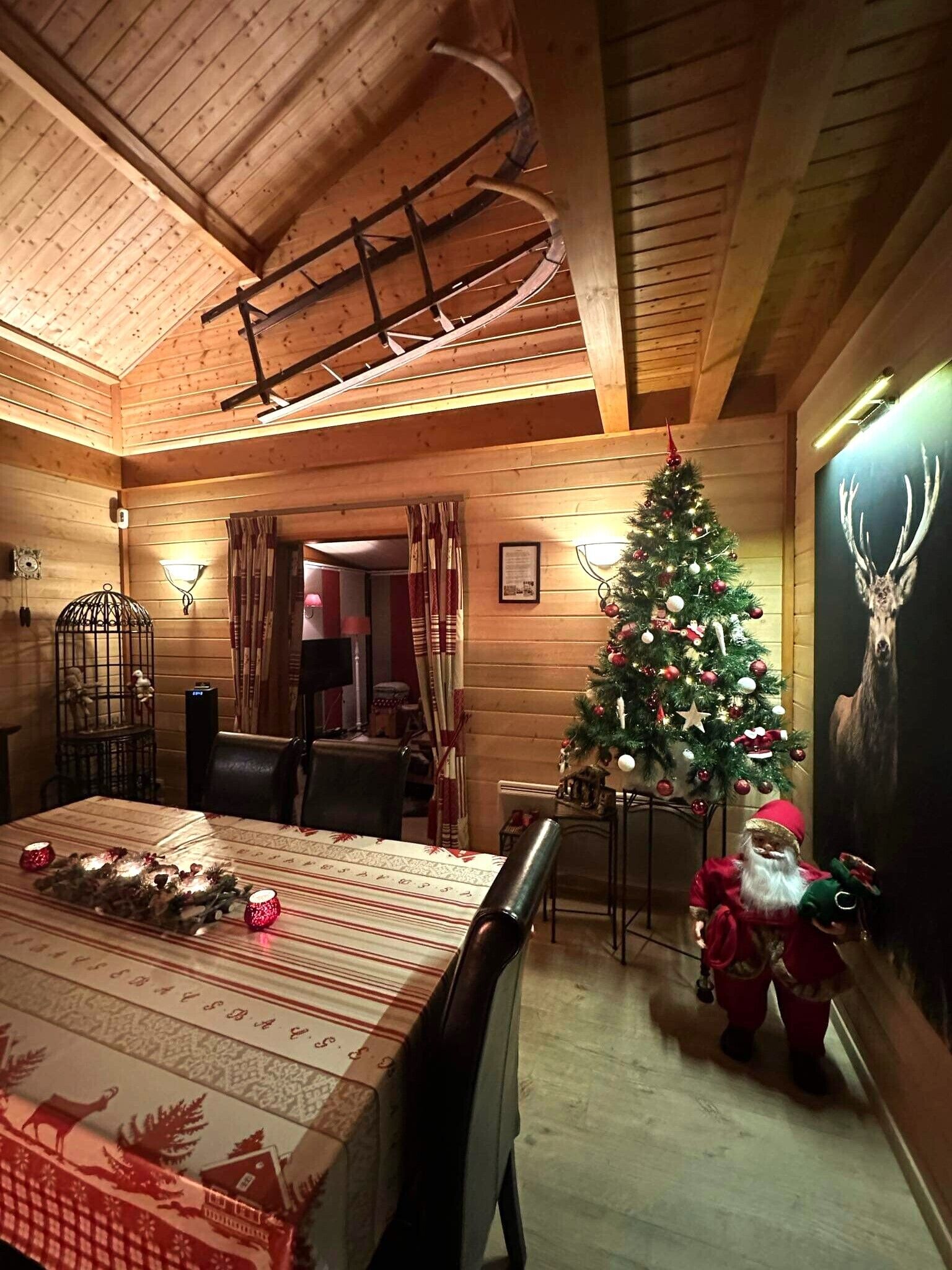 A cozy log cabin interior with a dining area, a decorated Christmas tree, and a wall-mounted deer poster.