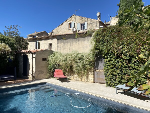 Une piscine avec un tuyau d’arrosage, un fauteuil de jardin rouge et un bâtiment en pierre doté d’un toit en tuiles et d’une antenne parabolique.
