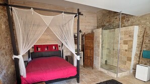 A bedroom with a four-poster bed, a red mattress, a black rug, and a shower area with glass doors. - Village house with heated pool in the Luberon for 14 people (Ansouis)