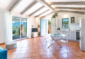 A spacious living area with a glass dining table, clear chairs, a flat-screen TV, and a kitchen with white cabinets and a window.