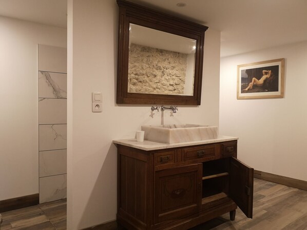 Un baño con lavamanos de mármol, tocador de madera, pared de piedra y un cuadro enmarcado.