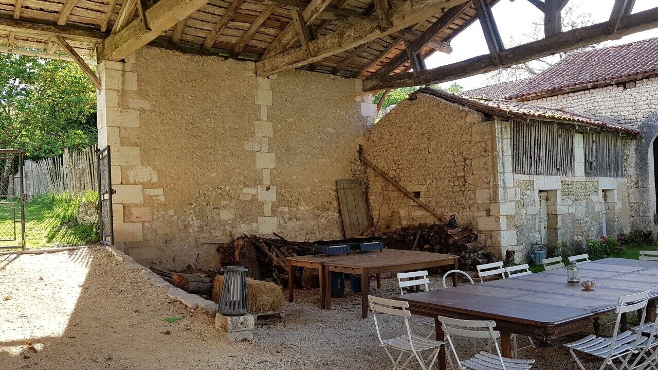 A rustic outdoor dining area with a wooden table and chairs, surrounded by stone walls and a thatched roof structure.