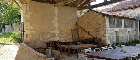 A rustic outdoor dining area with a wooden table and chairs, surrounded by stone walls and a thatched roof structure.