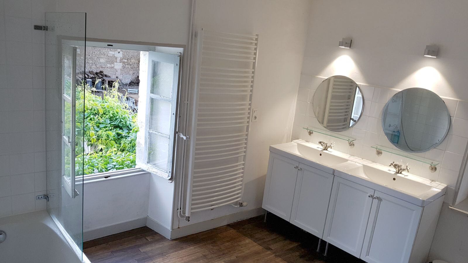 Une salle de bain avec un meuble-lavabo blanc, un miroir rond et une fenêtre donnant sur un paysage verdoyant.