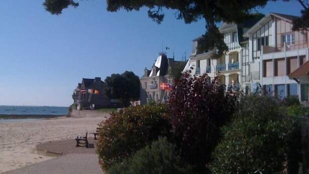 A beach with a sandy shore, a pathway leading to a building with multiple balconies, and a clear blue sky. - CHARMING HOUSE (Le Pouliguen)