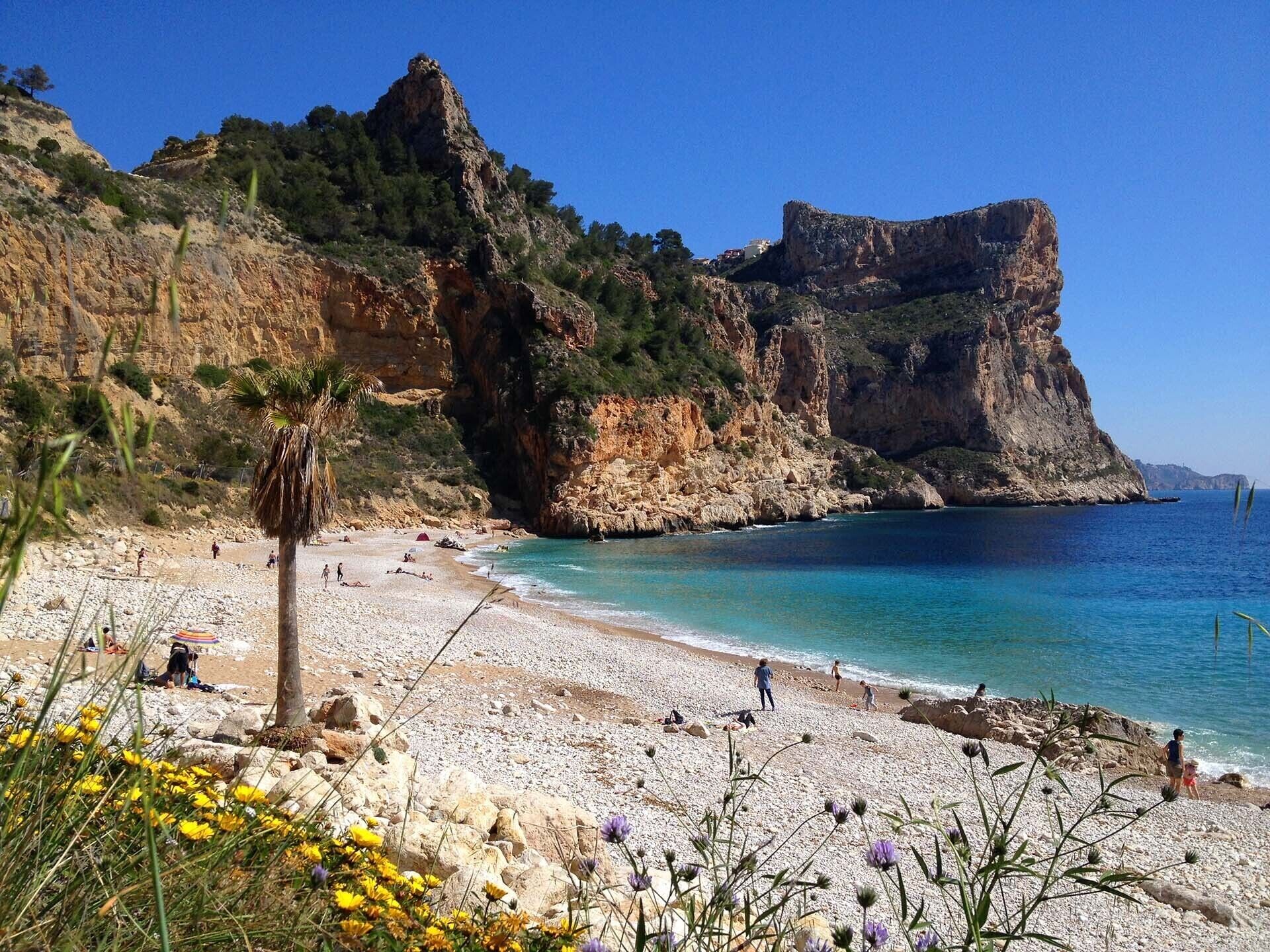 Une plage bordée de falaises rocheuses, avec une eau d’un bleu limpide, où les gens se prélassent au soleil.