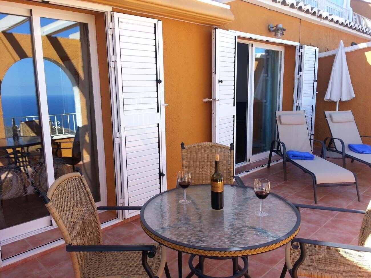 Un patio con una mesa redonda, dos sillas de mimbre, una botella de vino y una copa sobre la mesa.