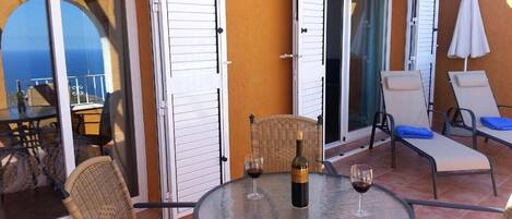 Un patio con una mesa redonda, dos sillas de mimbre, una botella de vino y una copa sobre la mesa.