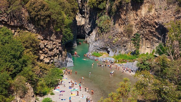 Paesaggio roccioso naturale con un fiume, scogliere e una spiaggia con persone e ombrelloni.