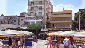 Una zona sulla spiaggia con diversi edifici, posti a sedere all'aperto sotto ombrelloni a righe e persone che passeggiano.