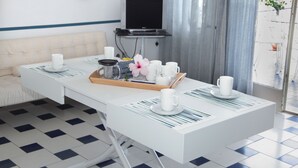 Una moderna camera d'albergo con un divano bianco, un tavolo da pranzo con un vassoio contenente tazze bianche e un fiore, e un televisore su un mobile.