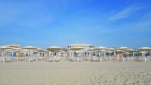 A beach with rows of sun umbrellas and lounge chairs, a building in the background, and clear skies. - Comfortable apartment in the center with air coditioning and sea view balcony (Lido di Pomposa)