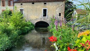 A rustic stone building with a small arched bridge over a narrow waterway, surrounded by lush greenery and vibrant flowers. - Superb 19th C. Farmhouse in extensive grounds, near Pyrenées, Pau and Lourdes (Caixon)