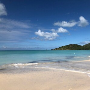 Une plage à l’eau turquoise et limpide, un rivage sablonneux et, au loin, une colline couverte de végétation.