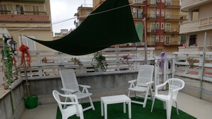 Eine Dachterrasse mit weißen Plastikstühlen und einem kleinen Tisch, umgeben von synthetischem Grünrasen.