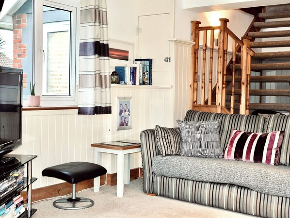 A living room with a striped sofa, a black leather ottoman, a small white table, and a wooden staircase.