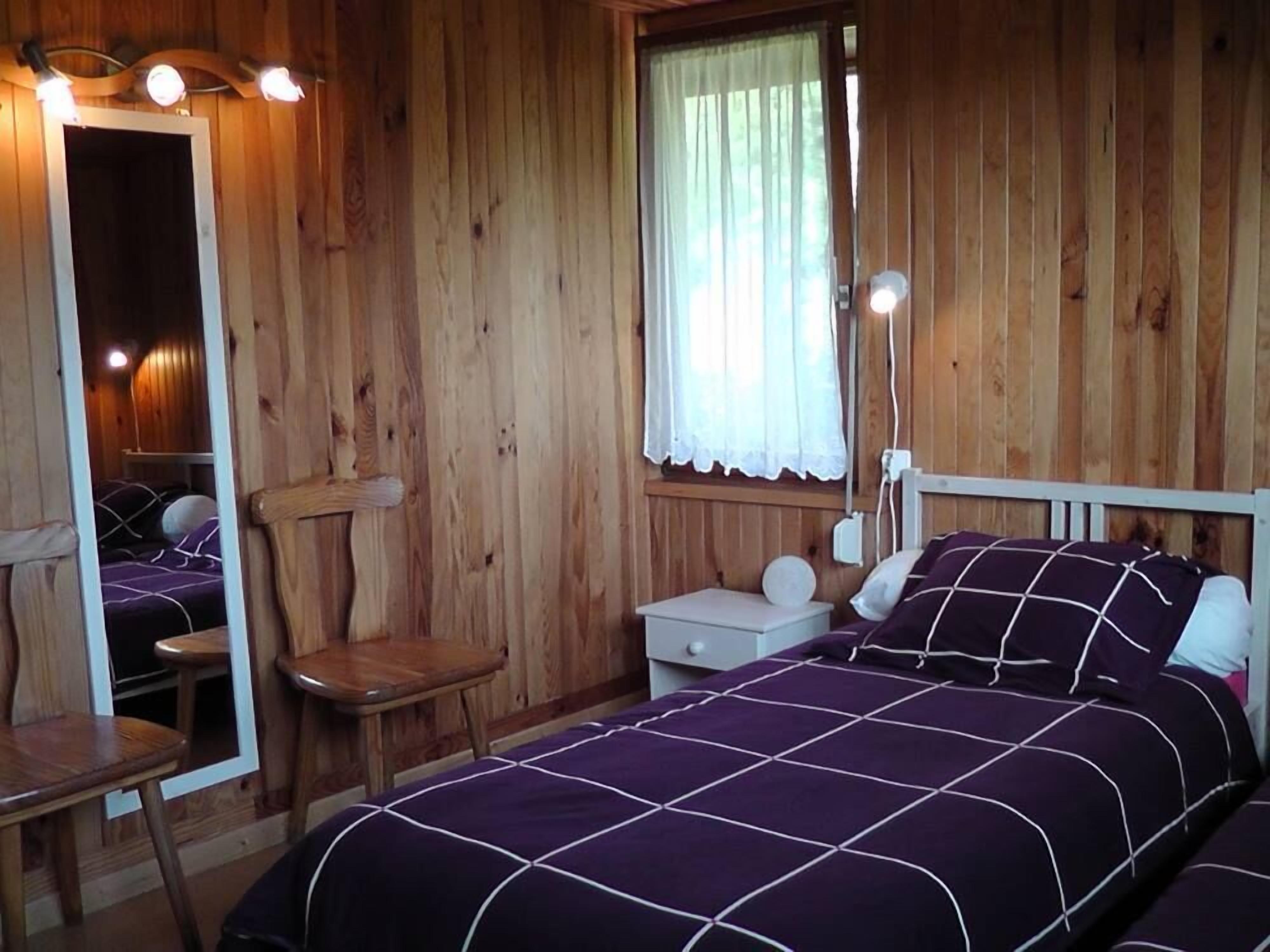 A wooden cabin room with a bed, a chair, a nightstand, and a mirror.