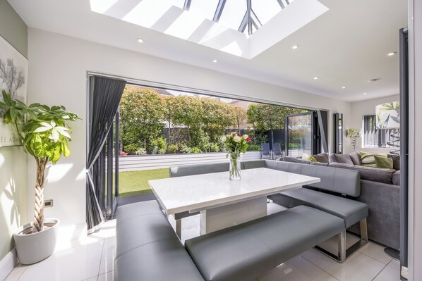 A modern interior with a white table, grey seating, and a large window offering a view of a garden.