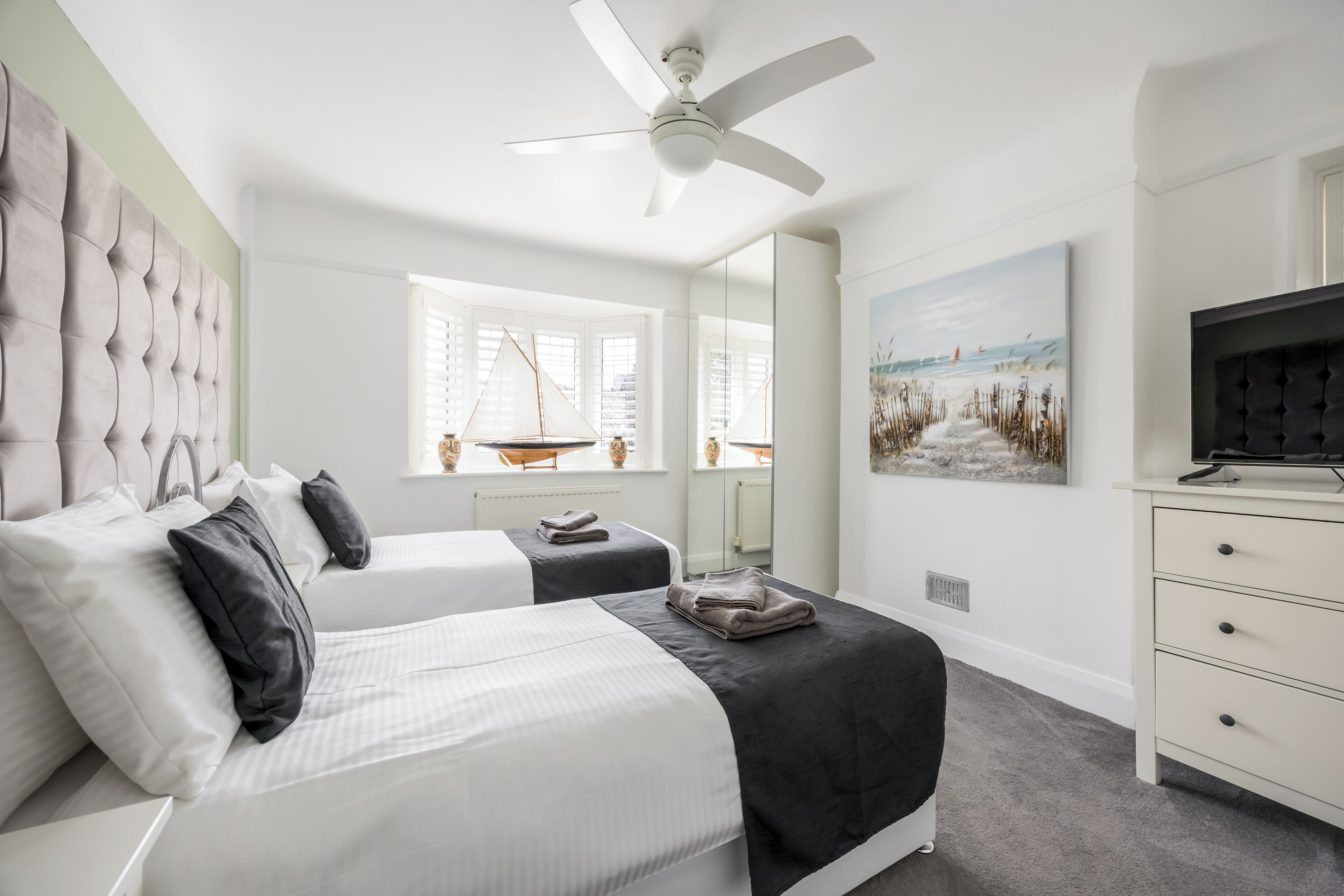 A bedroom with two beds, a ceiling fan, a large window with a view, a painting, and a TV on a dresser.