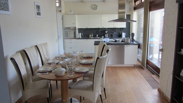 Un coin repas avec une table ronde dressée pour six personnes, entourée de six chaises. On aperçoit une cuisine en arrière-plan, avec des armoires blanches et des appareils électroménagers en acier inoxydable.