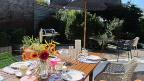 Un espace de restauration en plein air avec une table en bois dressée pour un repas, entourée de chaises et d’un parasol.