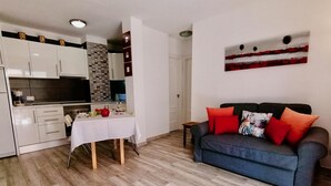A modern living room with a grey sofa and red cushions, a dining table with a white tablecloth, and a kitchen with white cabinets and a tiled backsplash. - Moon Holiday Home in Los Cristianos (Los Cristianos)