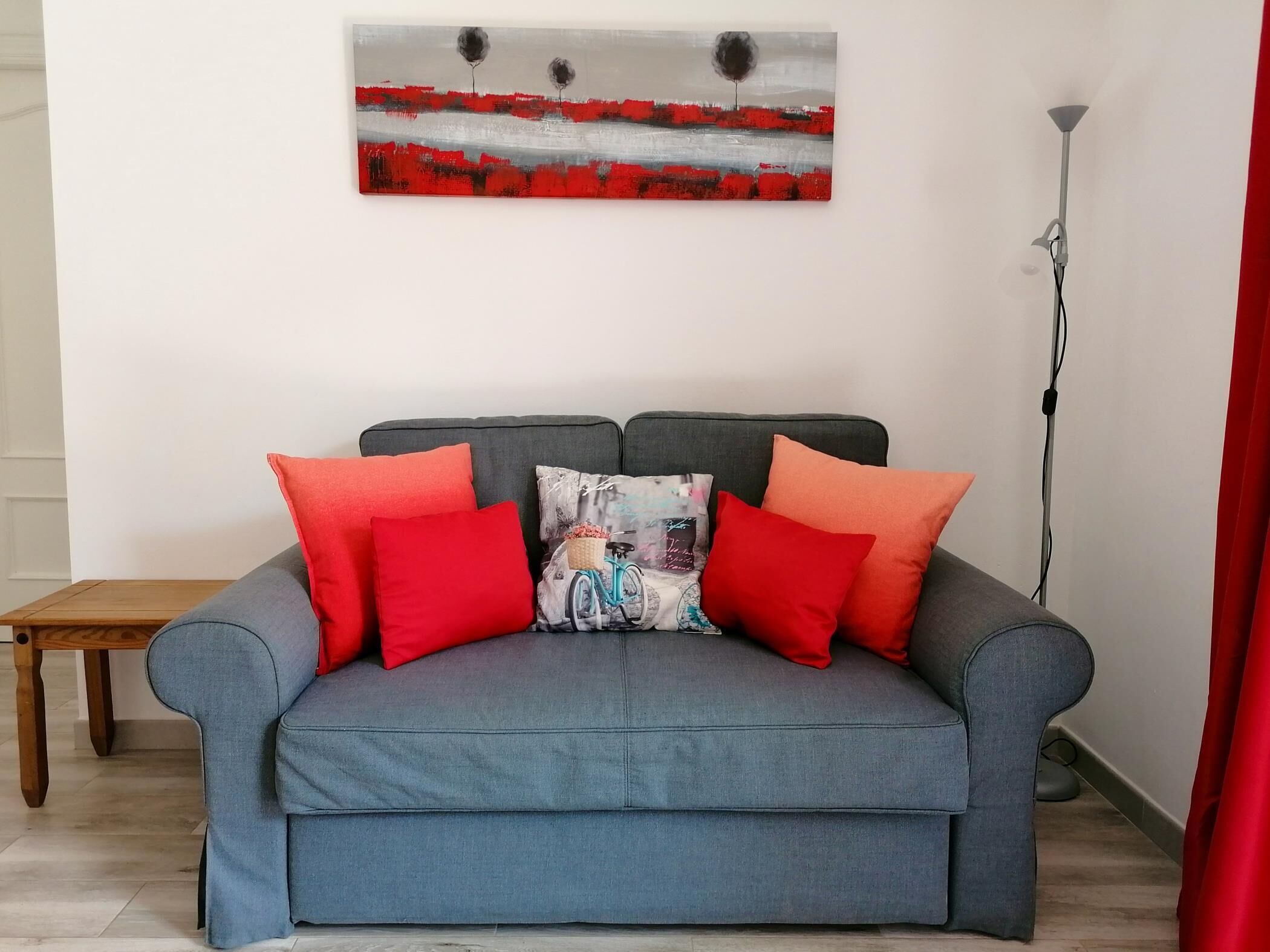 Ein graues Sofa mit roten und blauen Kissen, ein Holznachttisch und ein Gemälde an der Wand.