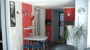 A dining area with a white table and blue chairs, a red accent wall, and a kitchen in the background. - The creeks, the sea, the hikes and a beautiful apartment with swimming pool. (Ensuès-La-Redonne)