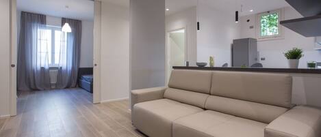 Una sala de estar moderna con un sofá, una zona de cocina con mostrador y electrodomésticos, y un pasillo que conduce a otra habitación.