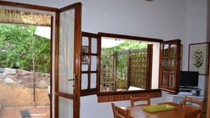 Uma sala de jantar com mesa e cadeiras de madeira, uma janela com vista para o verde e uma televisão sobre um móvel.