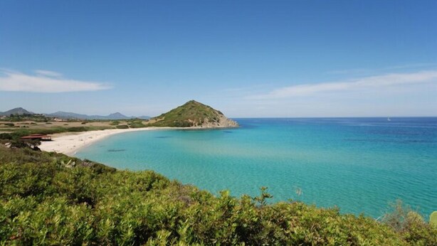 Paisagem costeira com uma praia de areia, águas turquesa cristalinas e uma pequena colina.
