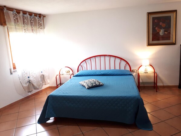 A bedroom with a red metal bed frame, a blue bedspread, two bedside tables with lamps, a window with sheer curtains, and a framed picture on the wall.